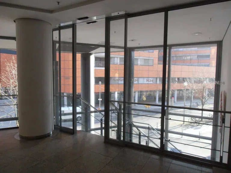 Eine verglaste Lobby mit großen Fenstern, die auf eine Straße und moderne Backsteinbauten blicken. Draußen sind Treppen und ein Geländer zu sehen, und durch die Fenster strömt Sonnenlicht herein.