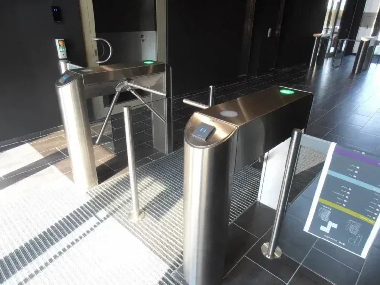 Zwei Drehkreuze aus rostfreiem Stahl mit Kartenlesegeräten kontrollieren den Zugang zu einer modernen Eingangshalle mit dunklen Fliesen und natürlichem Licht aus den nahe gelegenen Fenstern. Ein buntes Wegweiserschild ist teilweise auf der rechten Seite zu sehen.