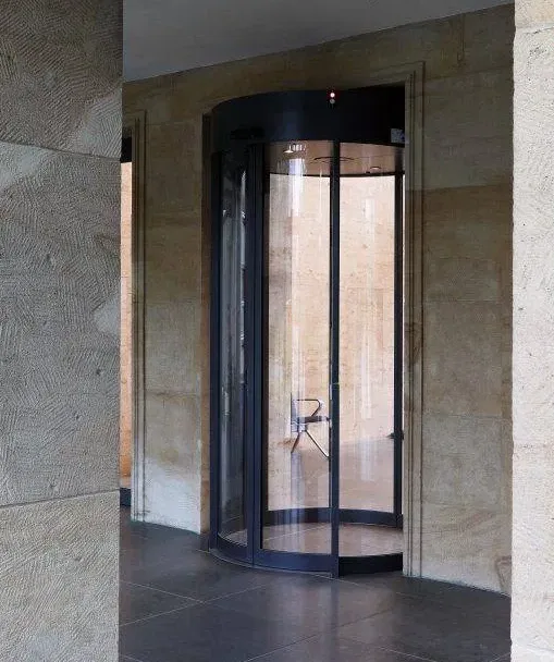 Ein einzelner Stuhl steht in einer gläsernen Drehtür in einem Gebäude mit Steinwänden und einem dunklen Fliesenboden. Die ungewöhnliche Platzierung des Stuhls verleiht der Szene ein surreales, leeres Gefühl.