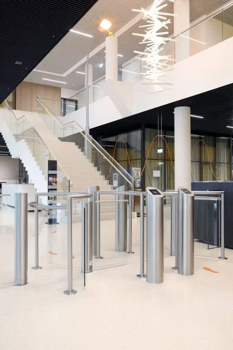 Ein modernes Foyer mit Drehkreuzen, einer breiten Treppe mit Glasgeländer und modernen Beleuchtungskörpern. Der Raum zeichnet sich durch schlichte, minimalistische Designelemente und einen polierten Boden aus.