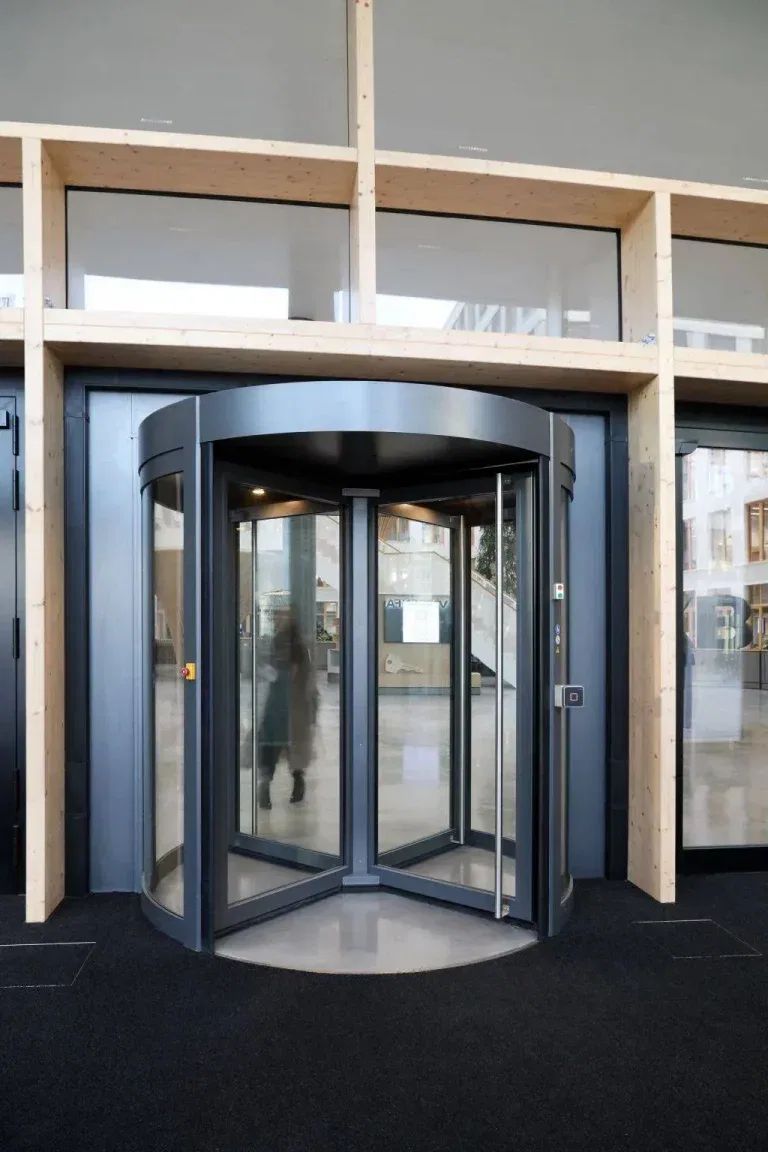 Eine moderne Karusselltür aus Glas mit schwarzem Rahmen ist im Eingang eines Gebäudes mit hellen Holzakzenten und großen Glasfenstern installiert. Der Innenraum ist durch die Tür sichtbar.