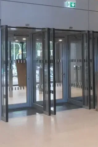 Eine Reihe von gläsernen Karusselltüren mit Metallrahmen in einer modernen Gebäudelobby, mit Spiegelungen auf dem Boden und Notausgangsschildern darüber.
