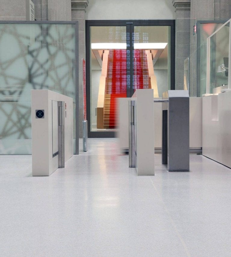 Ein modernes Gebäudeinneres mit Sicherheitsdrehkreuzen im Vordergrund, glatten hellen Fußböden, Glastrennwänden und einer breiten roten Treppe, die nach oben führt, im Hintergrund.